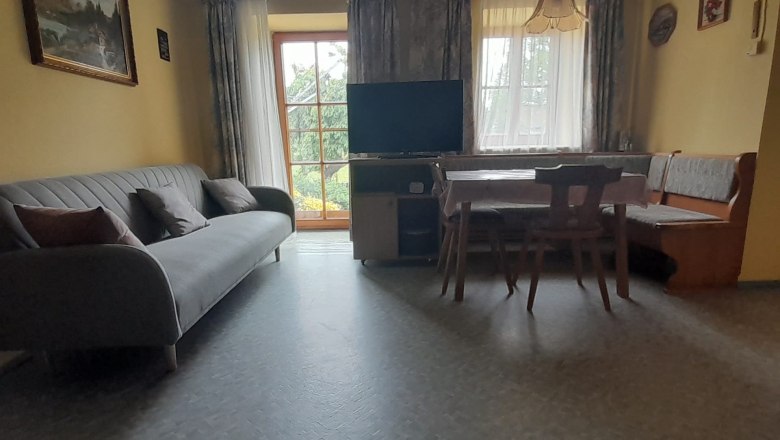 Wohnküche, © Erika Polt Wohnküche mit Sofa, Esstisch und Fernseher vor großen Fenstern.