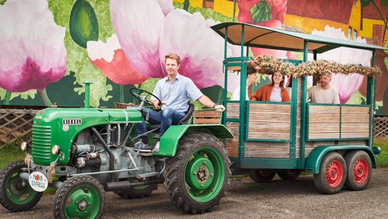 Mohnwirt Neuwiesinger, © Waldviertel Tourismus, Gerhard Wasserbauer Ein Mann fährt einen grünen Traktor mit Anhänger vor einer bunten Wand mit Mohnblumen.
