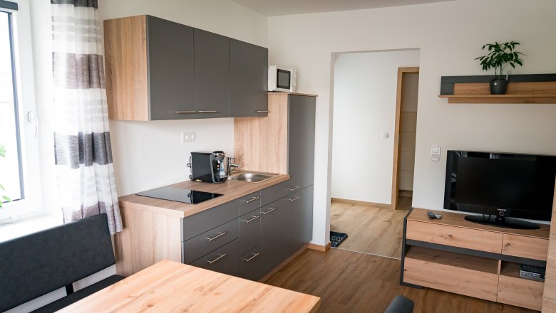 Living/dining room with view to the entrance, © Manuel Hahn Modern kitchen with gray cabinets, wooden worktop, coffee machine, sink, microwave and TV on wooden shelf.