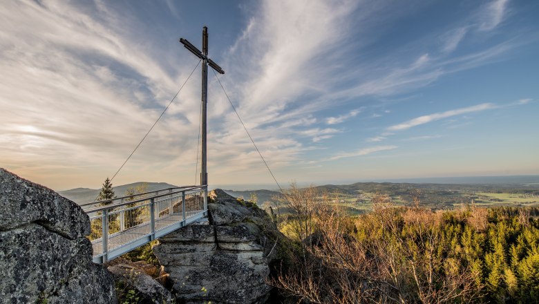 Ausflugsparadies Nebelstein, © Christian Freitag Ein Gipfelkreuz auf einem Felsen mit einer Aussichtsplattform, umgeben von Wald und einem weiten Blick über die Landschaft unter einem blauen Himmel.