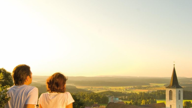 View Wachtstein, © Marktgemeinde Bad Traunstein Two people sit on a rock and look out over a church and a vast landscape at sunset.