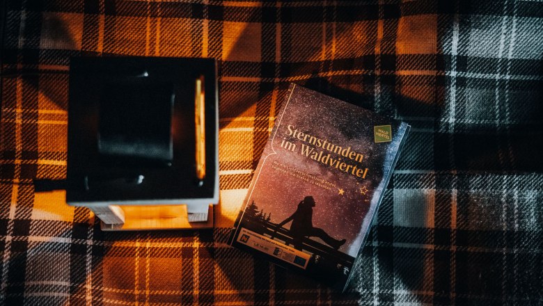 Waldviertler Sternengucker, © Waldviertel Tourismus, Line Sulzbacher Ein Buch mit dem Titel 'Sternstunden im Waldviertel' liegt auf einer karierten Decke neben einer Laterne.