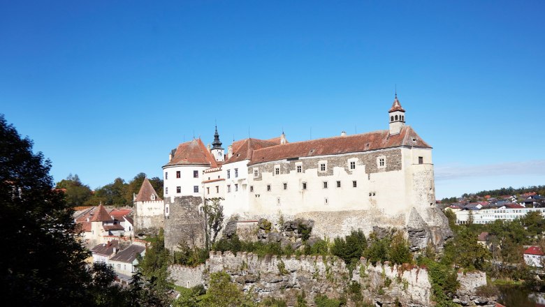 Burg Raabs, © Waldviertel Tourismus, lichtstark.com Burg Raabs auf einem Felsen über der Stadt, umgeben von Bäumen und blauem Himmel.