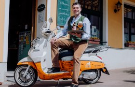 Biersommelier Andreas Frey, © Niederösterreich Werbung/thecreatingclick.com Ein Mann sitzt auf einem Roller vor einem Gebäude und hält ein Tablett mit Bierflaschen und einem Glas.