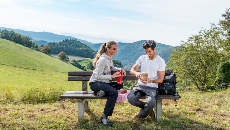 Wandern am Lebensweg, © Waldviertel Tourismus, Studio Kerschbaum Zwei Personen sitzen auf einer Bank in einer hügeligen Landschaft und machen eine Pause beim Wandern.