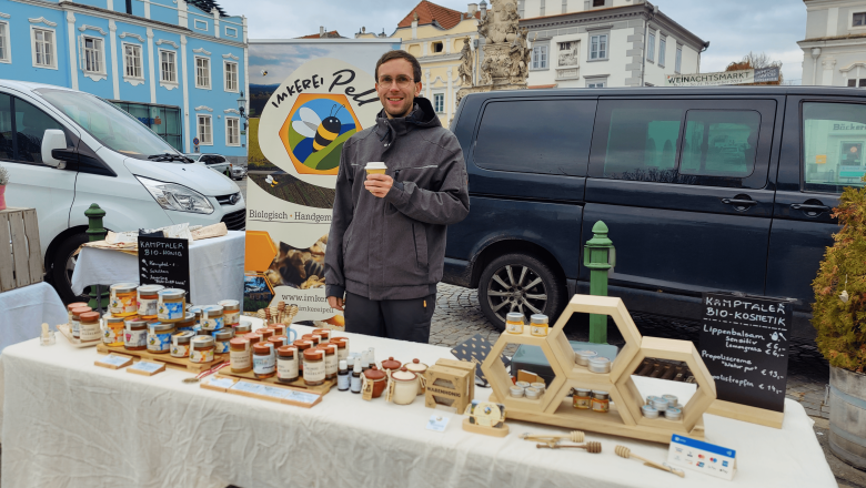 Alex am Markt in Langenlois, © Imkerei Pell Ein Mann steht an einem Marktstand mit Honigprodukten und hält einen Becher. Im Hintergrund sind ein Van und Gebäude zu sehen.