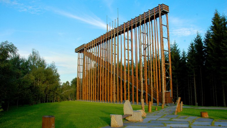 Himmelsleiter, © Preißl Eine hohe Holzkonstruktion im Freien, umgeben von Bäumen und grünem Gras.