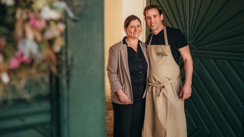 Landlords Isabelle and Philipp Wimmer-Joannidis, © Niederösterreich Werbung / Daniela Führer A couple stands smiling in front of a green wooden door, the man wearing an apron.