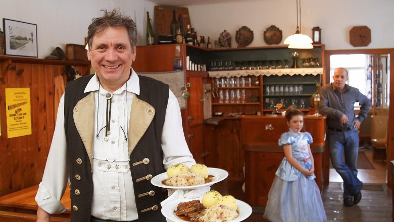 Schweinsbraten mit Waldviertler Erdäpfelknödel und Speckkrautsalat, © Fam. Mayrhofer, Wirtshaus zur Minidampfbahn Ein Mann in traditioneller Kleidung serviert Schweinsbraten mit Knödeln in einem rustikalen Gasthaus. Im Hintergrund ein Mann und ein Mädchen im Kleid.