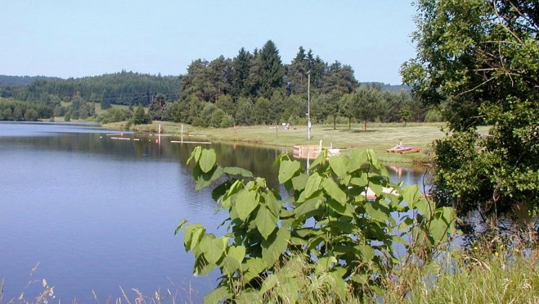 Badestrand Frauenwieserteich, © Gemeinde Langschlag Ein ruhiger See mit grünem Ufer und Bäumen im Hintergrund.