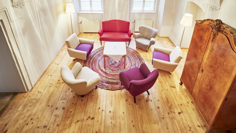 Haus Rosalia im Kloster Pernegg, © Schewig Dieter Ein gemütliches Teezimmer mit bunten Sesseln, einem roten Sofa und einem runden Teppich auf Holzboden. Ein Kronleuchter hängt von der gewölbten Decke.