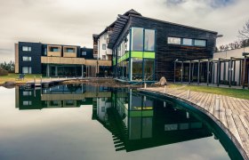 schwarzalm wellness, © Schwarz Alm Modernes Wellnesshotel mit Holzfassade und großem Teich im Vordergrund.