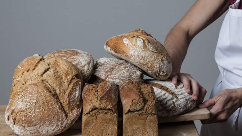 First Waldviertel organic baking school, © Erste Waldviertler Bio-Backschule, Andrea Peller Various loaves of bread on a wooden board, held by a person in an apron.