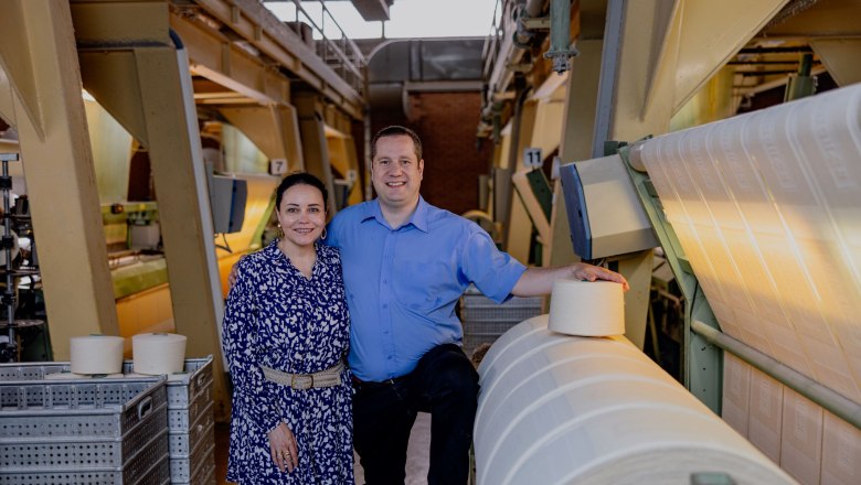 Katherine and Philipp Schulner, © Waldviertel Tourismus, Matthias Streibel Two people stand next to machines and rolls of fabric in a textile factory.