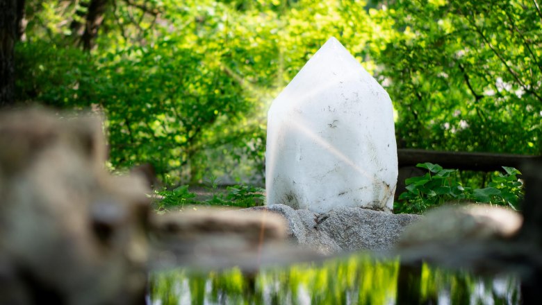 Amethyst World Maissau, © Amethyst Welt Maissau A large white crystal stands in a green forest, surrounded by plants and reflected in a small pond.