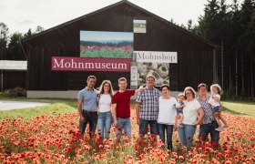 20230713karolinegrill_fam_gressl-hp, © Karoline Grill Eine Familie steht in einem Mohnfeld vor einem Gebäude mit der Aufschrift 'Mohnmuseum'.