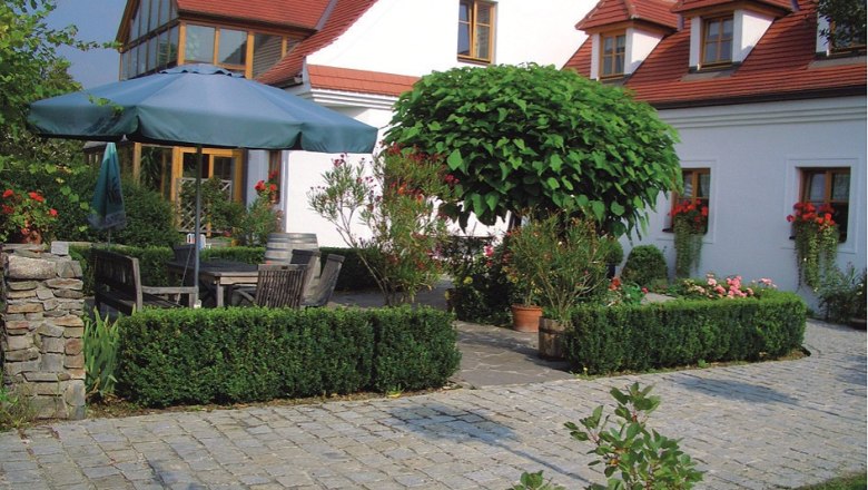 Winzergut Prabatsch-Aichinger, © Winzergut Prabatsch-Aichinger Ein gepflegter Garten mit Pflastersteinen, einem großen Sonnenschirm, Holzmöbeln und einem weißen Gebäude mit roten Dächern im Hintergrund.