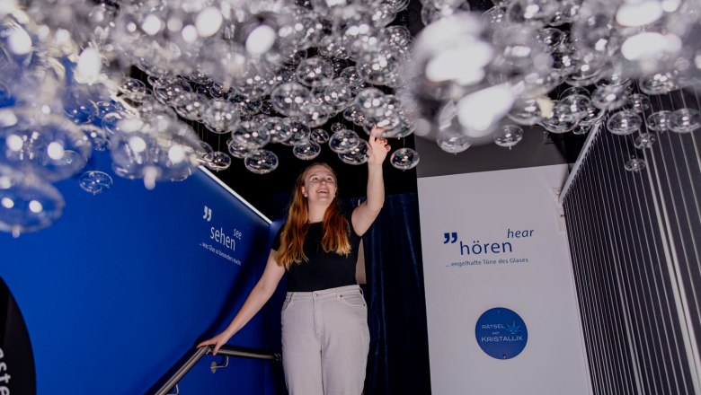 KRISTALLIUM glas.erleben., © Waldviertel Tourismus, Matthias Streibel Eine Frau berührt hängende Glasblasen in einer Ausstellung mit blauen Wänden und Beschriftungen.