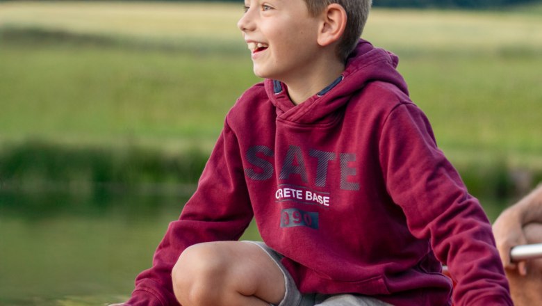 Bootsfahrt, © Familie Moser Ein Junge in einem roten Hoodie sitzt lächelnd auf einem Boot auf einem Teich..