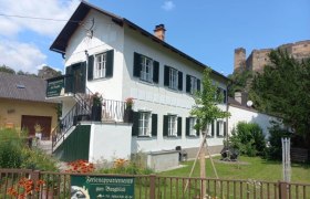 Appartements Zum Burgblick, © Jürgen Mokesch Ein weißes Gebäude mit grünen Fensterläden, umgeben von einem Garten, mit einer Burg im Hintergrund.