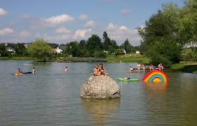 Obere Ortsteich Ottenschlag, © Gemeinde Ottenschlag Kinder spielen im Wasser eines Teichs mit bunten Schwimmreifen und einem großen Stein.
