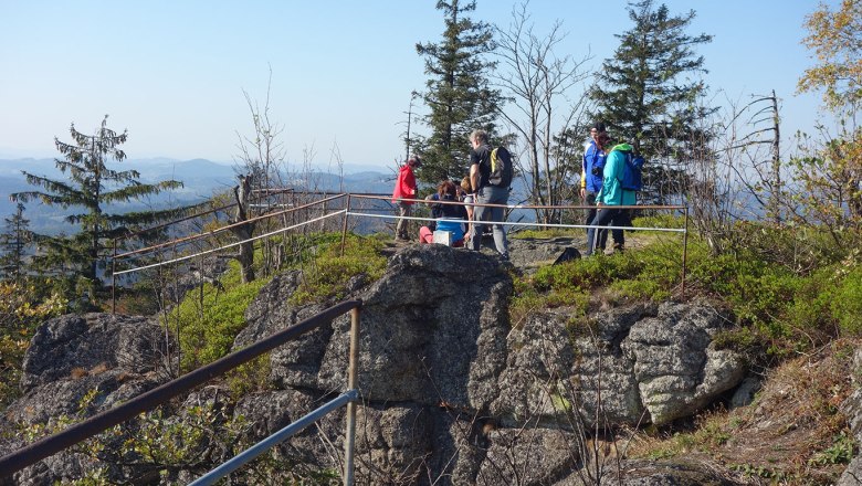 Aussichtsberg Burgsteinmauer, © Leo Baumberger Gruppe von Wanderern auf einem felsigen Aussichtspunkt mit Geländer, umgeben von Bäumen und Bergen im Hintergrund.