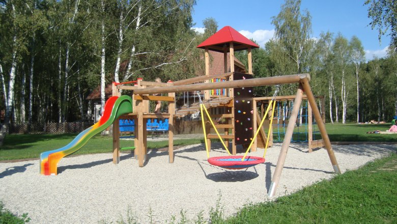 Children's playground, © Hannes Messmann A children's playground with slide, swings and climbing frame, surrounded by trees.