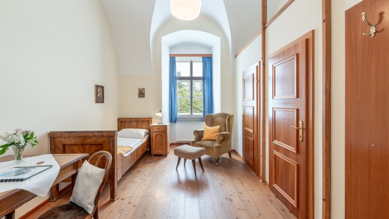 Stift Altenburg Einzelzimmer, © Martin Mathes Einzelzimmer im Stift Altenburg mit Bett, Sessel, Tisch und Fenster mit blauen Vorhängen.