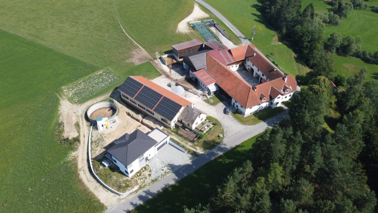 Knappenhof Hahn - Flugaufnahme, © Familie Hahn Luftaufnahme eines Bauernhofs mit mehreren Gebäuden und Solarpanelen auf einem Dach, umgeben von Feldern und Wäldern.