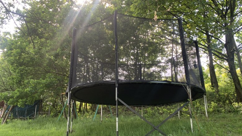 Spielwiese, © Sabine Kerschbaumer Ein Trampolin mit Sicherheitsnetz steht auf einer Wiese, umgeben von Bäumen. Sonnenstrahlen scheinen durch das Blätterdach.