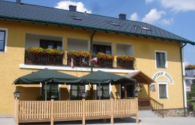 Gasthof Hanni KG, © Gasthof Hanni KG Gelbes Gebäude mit Balkon, Blumen und Terrasse mit Sonnenschirmen.