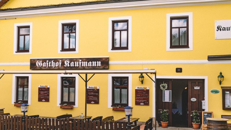Inn in Litschau, © Niederösterreich Werbung/Mara Hohla Yellow building with the inscription 'Gasthof Kaufmann', wooden tables and chairs outside.