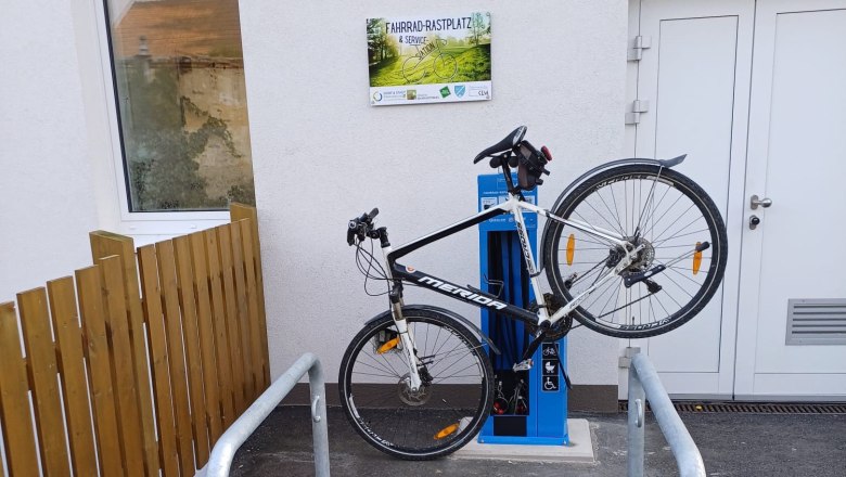 Fahrrad Rastplatz & Service-Station, © Gemeinde Meiseldorf Fahrrad an einer blauen Service-Station befestigt, neben einem Gebäude mit einem Schild 'Fahrrad-Rastplatz & Service Station'.