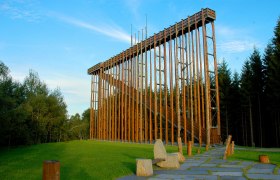 Himmelsleiter, © Preißl Eine hohe Holzkonstruktion im Freien, umgeben von Bäumen und grünem Gras.