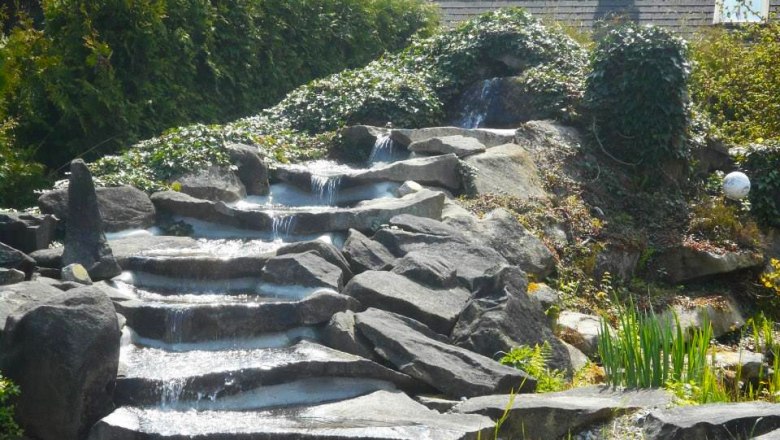Garden of 100 roses__ B.Scherzer, © Garten der 100 Rosen, B.Scherzer Stone waterfall in a garden with plants and bushes.