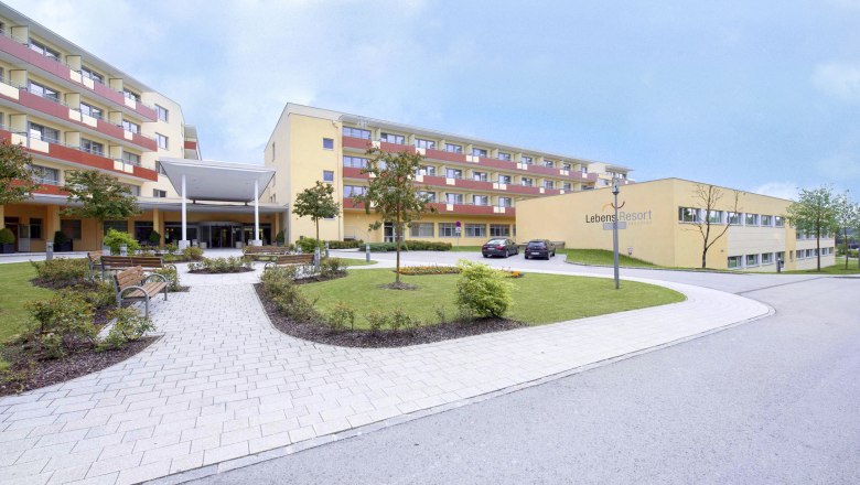 Hausansicht, © Xundheitswelt Außenansicht eines modernen Gebäudekomplexes mit gepflegtem Garten und Parkplätzen.