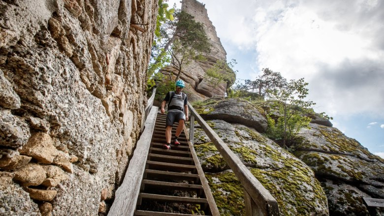 Arbesbach castle ruins, © Waldviertel Tourismus, Erwin Haiden A man in a helmet walks down a wooden staircase, next to a stone castle ruin and rocks.