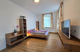 Apartment 2 - Schlafbereich, © Apartment Bauer Zur Alten Schule Modernes Schlafzimmer mit Holzboden, Doppelbett, Fernseher und Regalen. Große Fenster mit gestreiften Vorhängen.