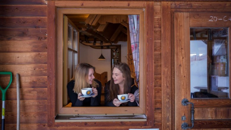 Kirchbochstadl, © Waldviertel Tourismus, Robert Herbst Zwei Frauen schauen aus einem Fenster einer Holzhütte und halten Tassen in den Händen.