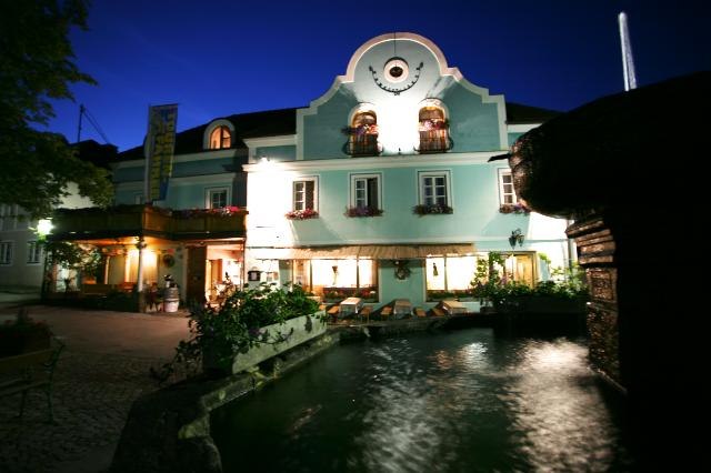 Inn-Pension "Drei Hacken", © starkmann A traditional inn with a blue façade and illuminated windows at night, with a fountain in the foreground.