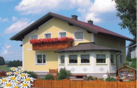 Haus Gertrud, © Fam. Sommer-Leitner Ein gelbes Haus mit rotem Dach und Blumenkasten auf dem Balkon, umgeben von einem Holzzaun und Gänseblümchen im Vordergrund.