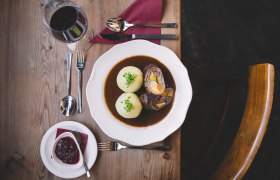 Rindsroulade mit Erdäpfelknödeln, © Niederösterreich Werbung/Mara Hohla Ein Teller mit Rindsroulade und zwei Erdäpfelknödeln in Soße, daneben ein Glas Rotwein und Preiselbeeren auf einem Holztisch.