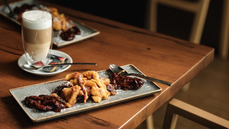 W4, © Weinviertel Tourismus / Michael Reidinger Ein Teller mit Kaiserschmarrn und Pflaumenkompott auf einem Holztisch, daneben ein Glas Latte Macchiato.
