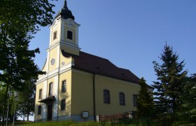 Pfarrkirche Haugschlag, © Gemeinde Haugschlag Pfarrkirche Haugschlag mit gelber Fassade und Turm, umgeben von Bäumen.