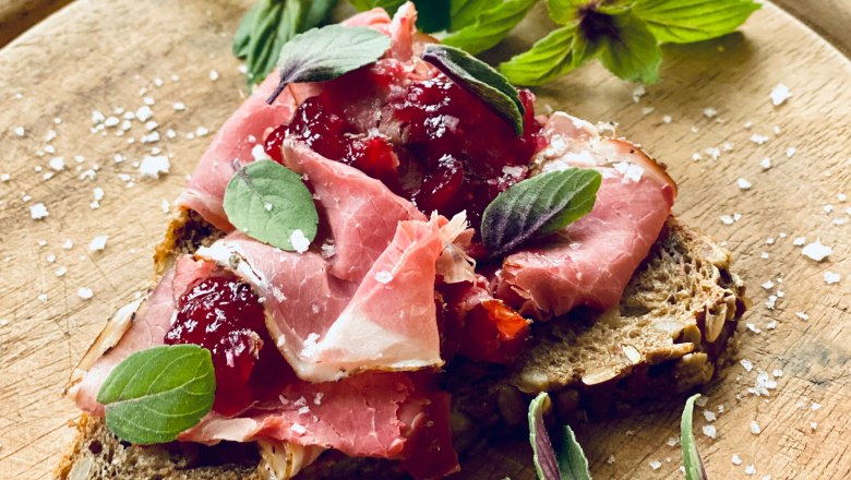 Wild-Geselchtes, © Amelie Seilern-Aspang Nahaufnahme eines belegten Brotes mit Schinken, Preiselbeeren und Kräutern auf einem Holzbrett.