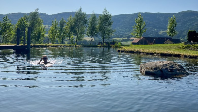 Schwimmen, © Familie Moser Person schwimmt in einem Teich mit Bäumen und Hügeln im Hintergrund.