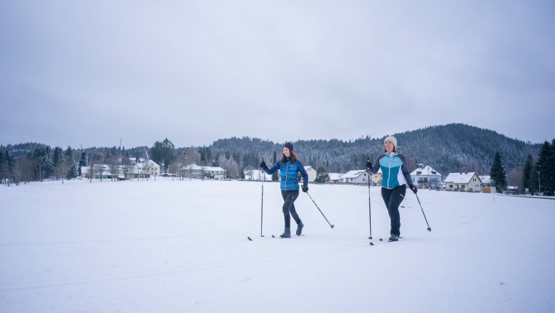 Langlaufen in Karlstift, © Waldviertel Tourismus, Robert Herbst Zwei Personen beim Langlaufen auf einer schneebedeckten Fläche vor einem Dorf und bewaldeten Hügeln.