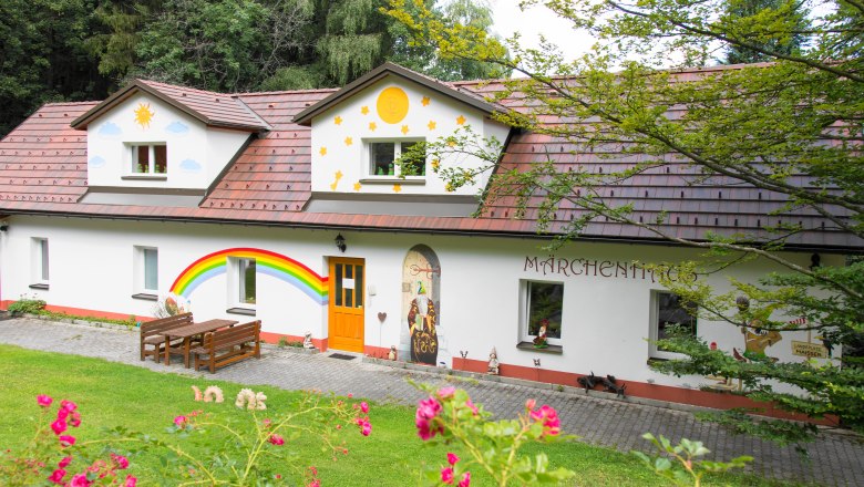 The fairytale house exhibition, © Andreas Schmidt A colorfully painted house with fairytale motifs and a rainbow on the wall, surrounded by green nature.