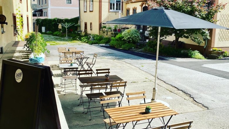 Gastgarten, © Hopfenspinnerei Ein Gastgarten mit Holztischen und Stühlen, einem Sonnenschirm und Pflanzen in einem Innenhof.