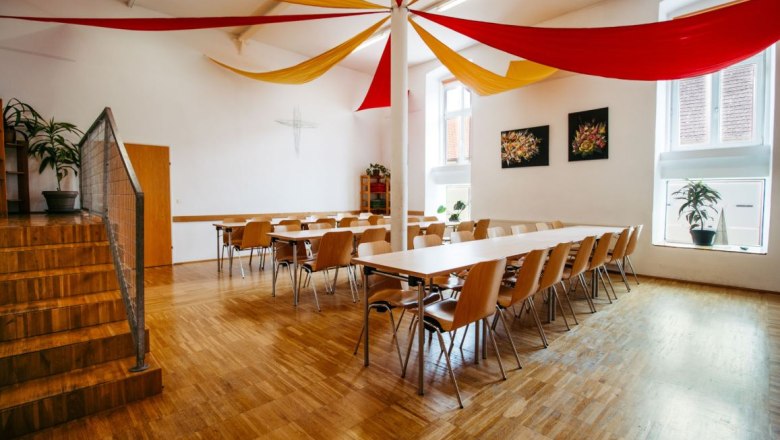 Dining room, © Melanie Köberl / Agnes Stefanie Wagner A bright dining room with a wooden floor, tables and chairs. Red and yellow fabric panels hang from the ceiling.